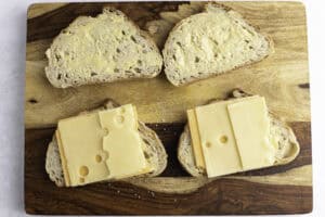 Slices of cheddar cheese and swiss cheese atop slices of buttered sourdough bread on a wooden cutting board.