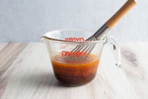 Side view of a glass measuring cup with mesquite marinade and a wooden-handled whisk.
