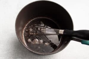 Overhead view of a black saucepan with tart cherry juice, lemon juice, lime juice, honey, and a whisk on a white countertop.