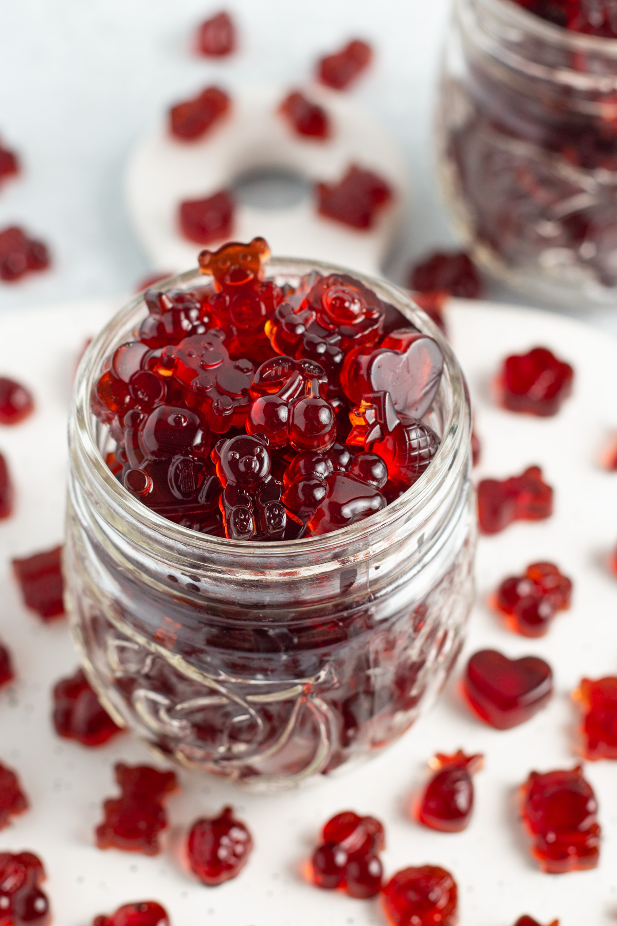 Close-up, overhead, angled view of a glass jar overflowing with bright red tart cherry gummies on a white countertop.