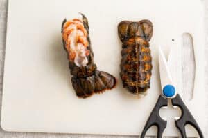 Overhead view of 2 lobster tails on a white cutting board, showing how to pull lobster meat out of one tail.