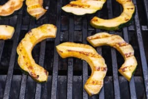 Overhead view of 3 acorn squash slices on a grill grate. Squash has dark grill marks seared into the flesh.