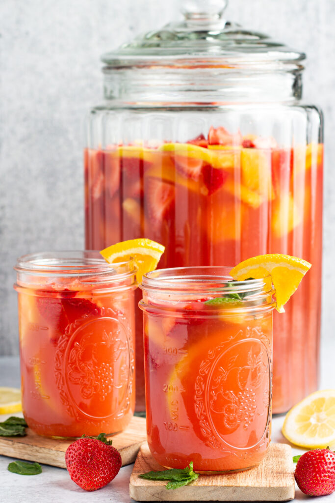 Side view of a large glass container of bright red jungle juice behind 2 small mason jars of jungle juice garnished with fruit and a clear straw.