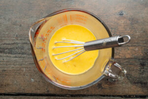 Overhead view of scrambled eggs and milk in a glass measuring cup with a silver-handled whisk.