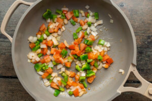 Overhead view of a large skillet containing sautéed onions, green bell peppers, and roma tomatoes.