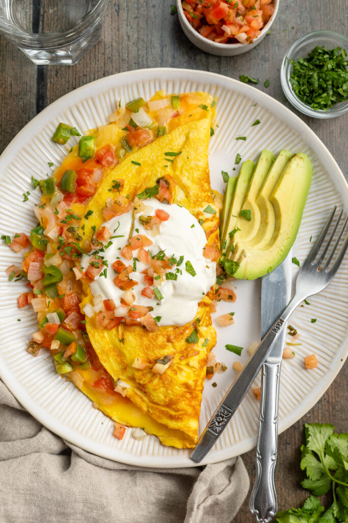Overhead view of a Mexican omelette with cheese, bell pepper, and onion, topped with sour cream and sliced avocado on a white plate.