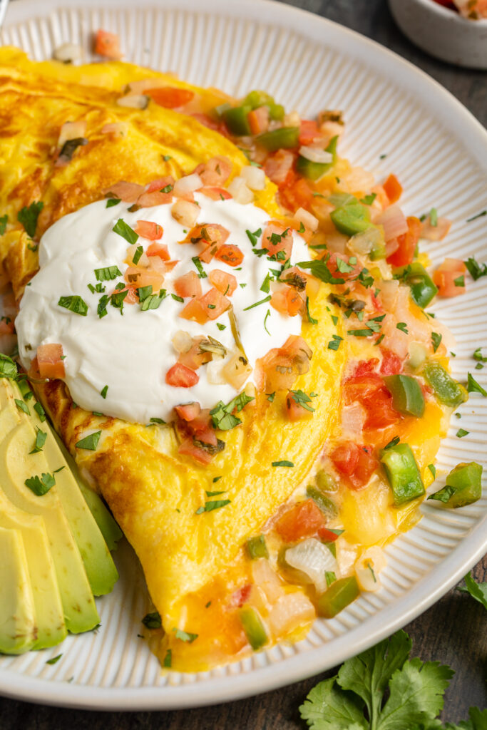 Angled view of a Mexican omelette filled with cheese, onion, and bell pepper, and topped with sour cream and slices of avocado.