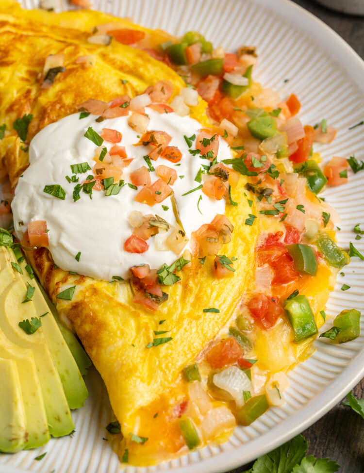 Angled view of a Mexican omelette filled with cheese, onion, and bell pepper, and topped with sour cream and slices of avocado.