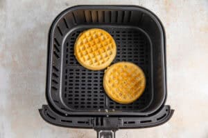 Overhead view of two round frozen waffles in an air fryer basket.