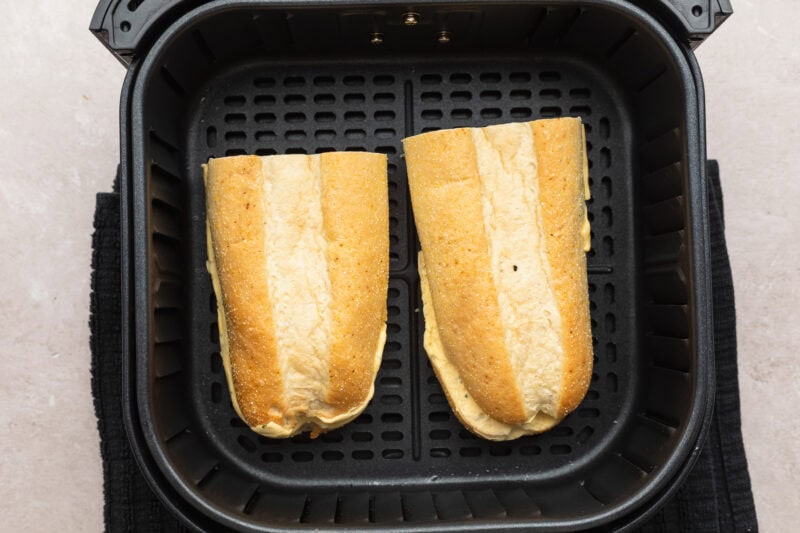 Overhead view of two halves of a frozen garlic bread loaf in an air fryer basket, before air frying.