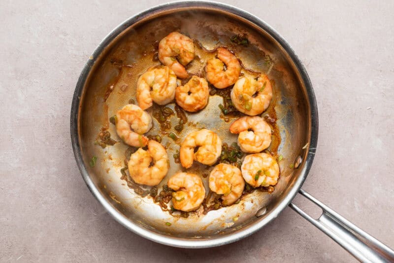 Overhead view of sautéed shrimp and diced green onion in a large silver skillet on a neutral countertop.