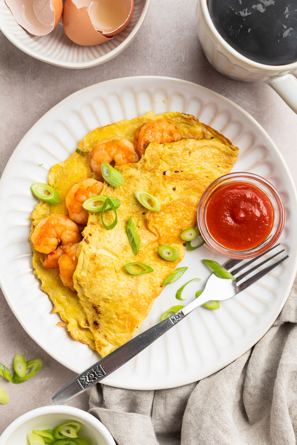 Overhead view of a folded, half-moon shaped shrimp omelette topped with chopped green onions on a white plate with small bowl of ketchup.