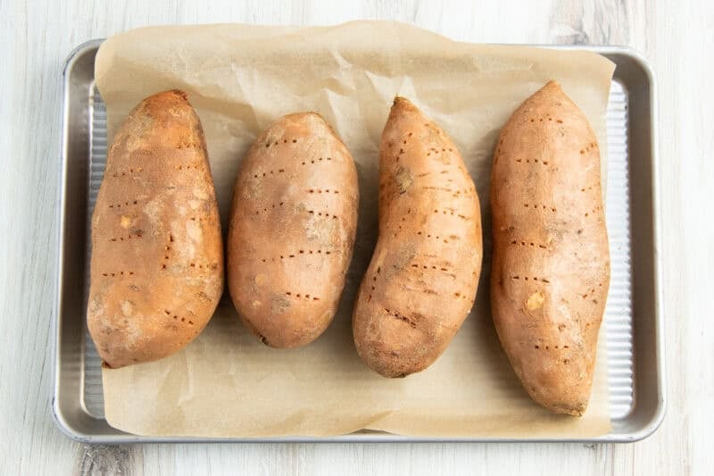 Overhead view of 4 large sweet potatoes lined horizontally on a baking sheet lined with parchment paper.