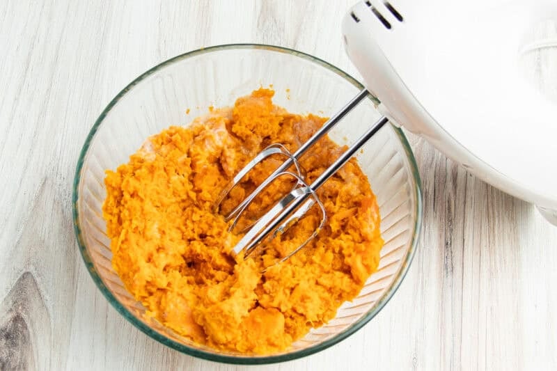 Overhead view of whipped sweet potatoes in a large glass mixing bowl with a white hand mixer fitted with beater attachments.