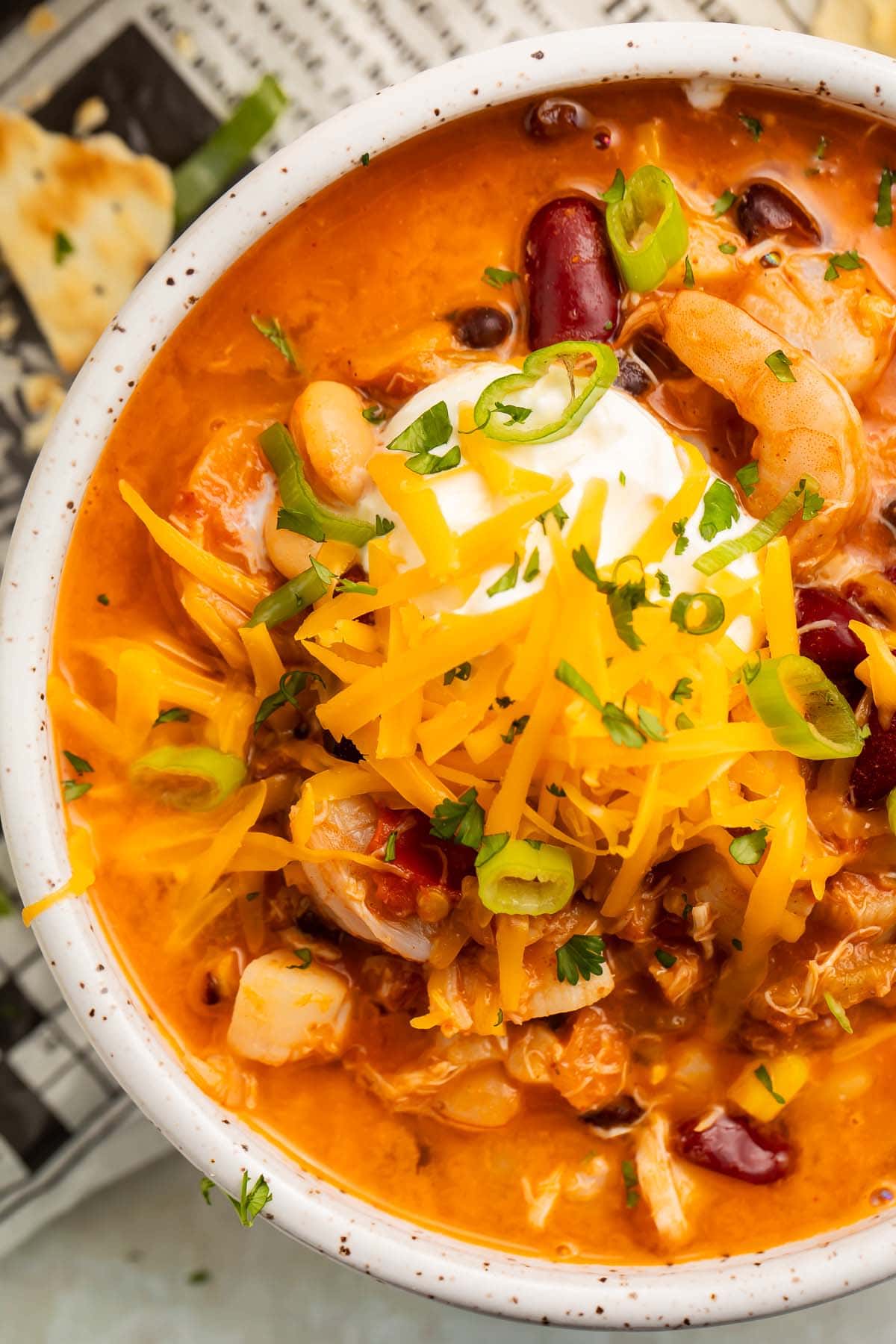 A large bowl of rich seafood chili, with shrimp and beans topped with sour cream, shredded cheddar, and sliced green onions.