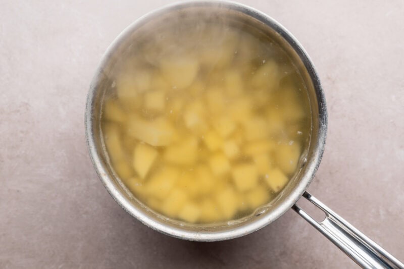 Overhead view of a silver pot holding boiling water, cubed potatoes, and salt on a neutral countertop.