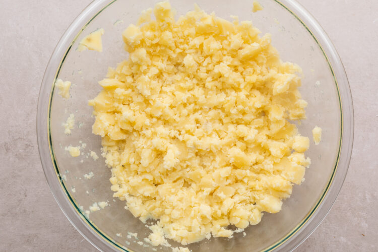 Overhead view of lightly mashed potatoes in a large glass mixing bowl on a neutral countertop.