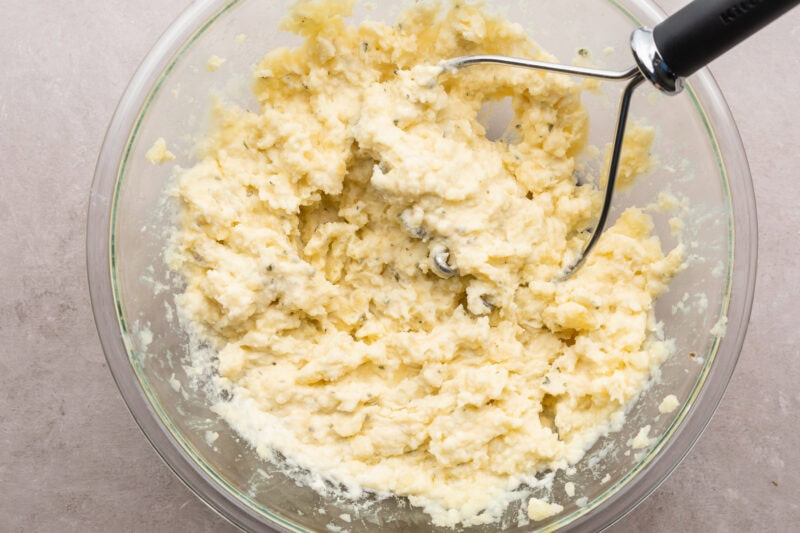 Overhead view of creamy mashed potatoes in a large glass mixing bowl with a potato masher resting on a neutral countertop.