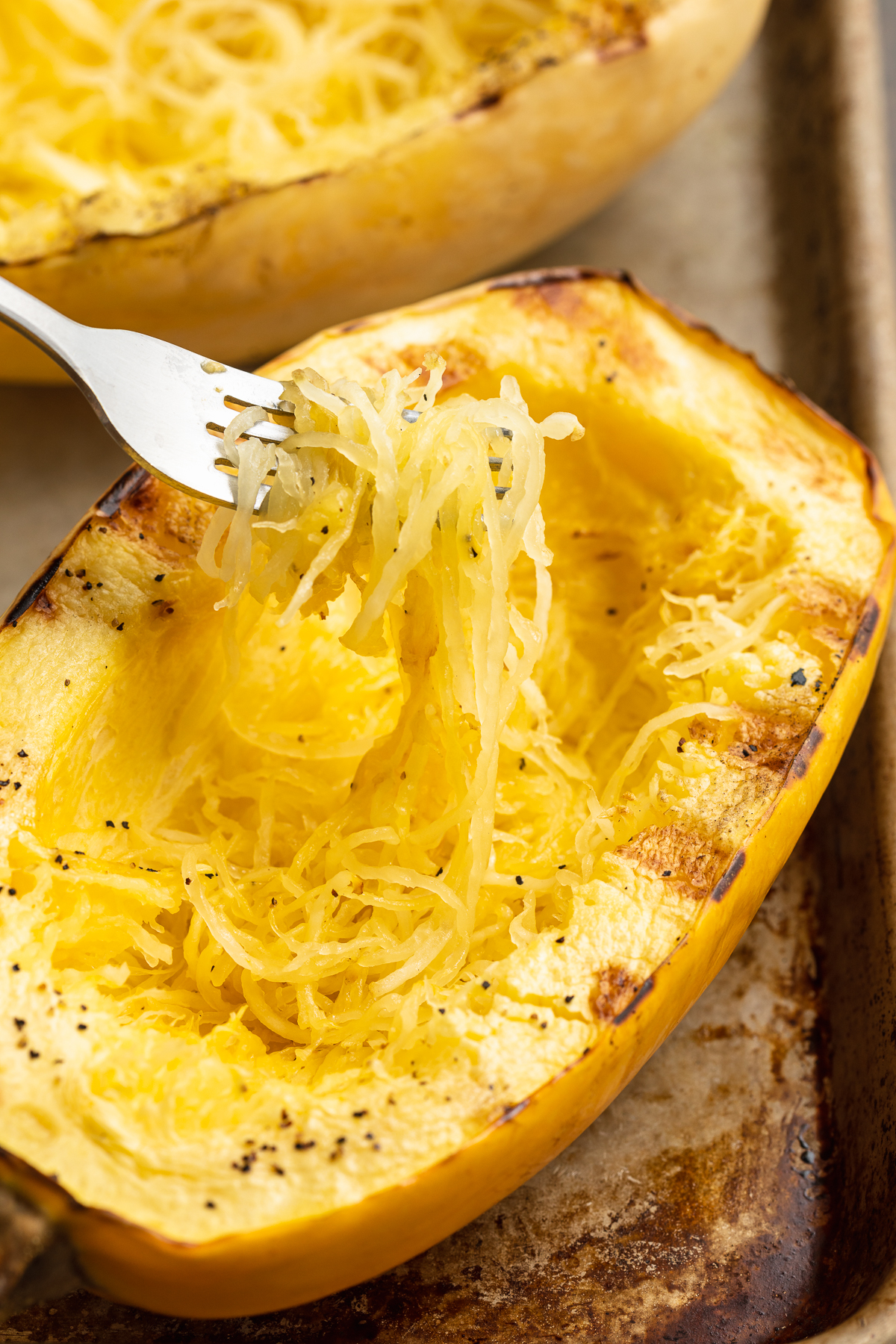 A fork lifts spaghetti squash out of a grilled half resting on a baking sheet.