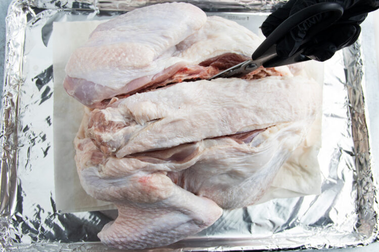 Overhead view of a hand covered by a black glove cutting into a raw, whole turkey to remove the backbone of the bird.