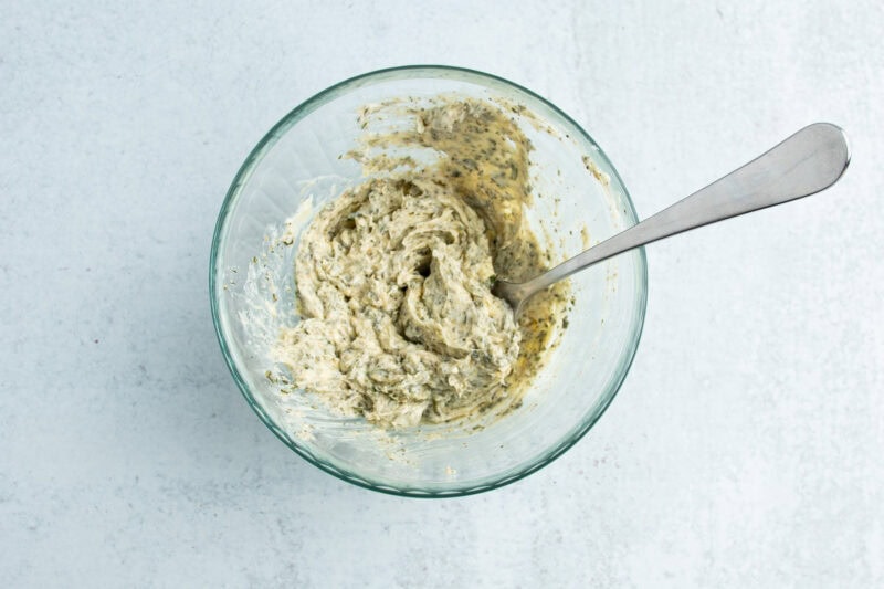 Overhead view of a small glass mixing bowl holding a compound butter mixture of garlic and fresh herbs.