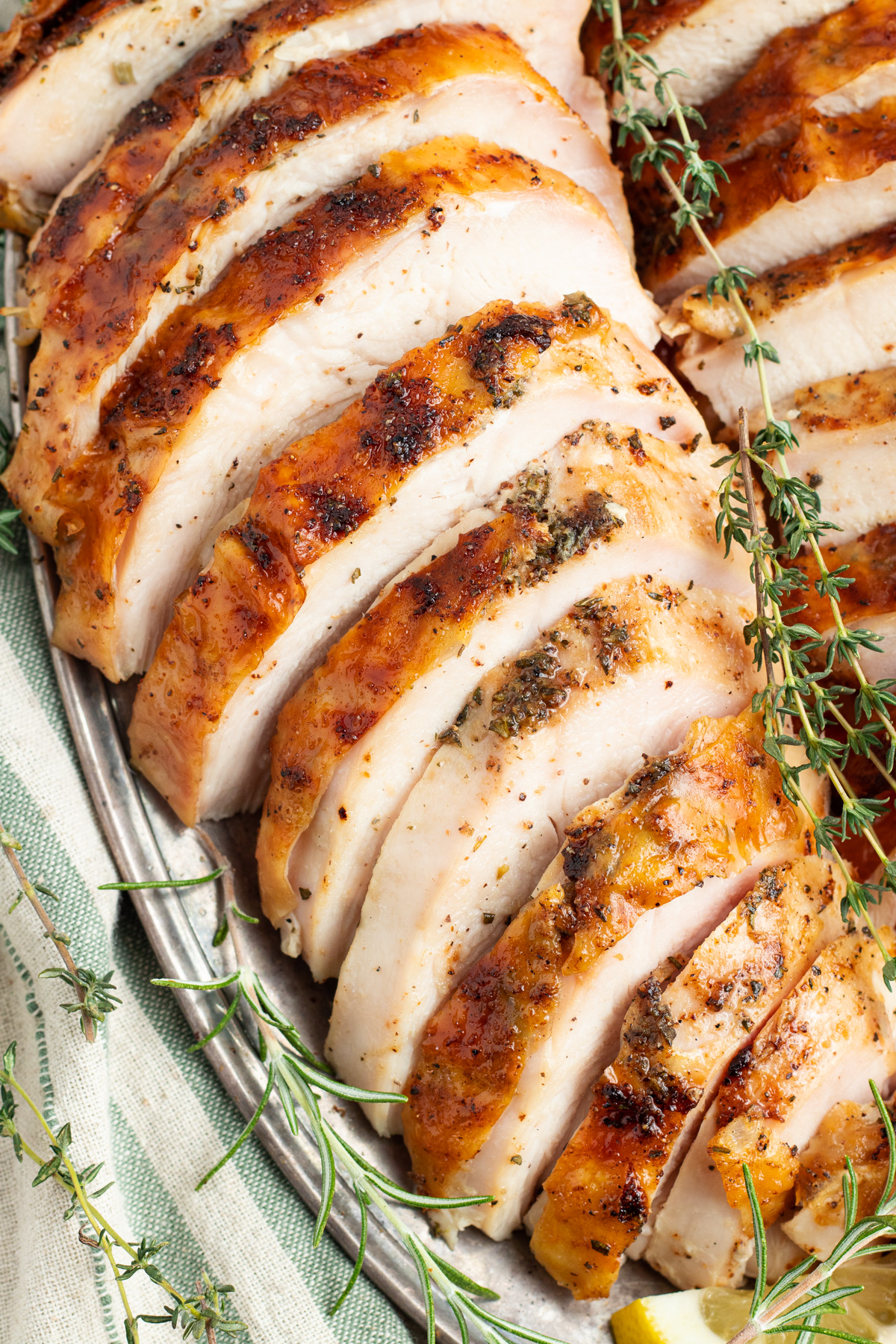 Close-up, overhead angled view of a thickly sliced grilled turkey breast on a platter with sprigs of green herbs.