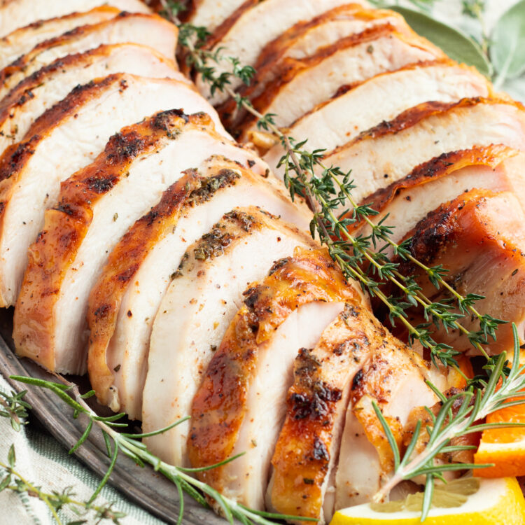 Overhead, zoomed in view of grilled turkey breast cut into thick slices and arranged on a serving platter with fresh herbs and lemon wedges.