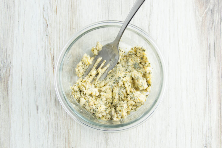 Overhead view of a small glass mixing bowl holding a mixture of butter, herbs, and seasonings with the fork used for mixing.