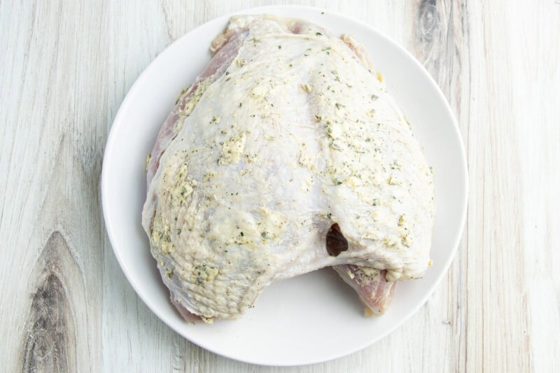 Overhead view of a brined turkey breast rubbed with compound-butter on a round white plate.