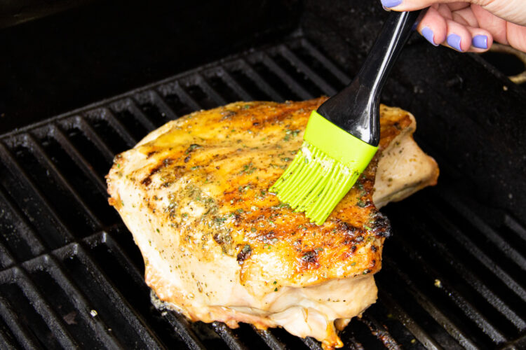 A view of a turkey breast on a grill while a hand holding a basting brush brushes additional butter over grilled skin.