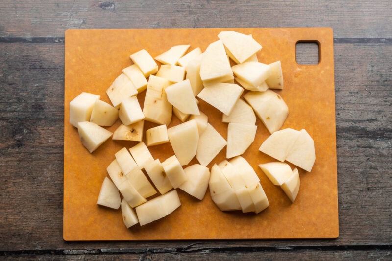 Overhead view of peeled, chopped potatoes on a wooden cutting board resting on a dark wooden tabletop.