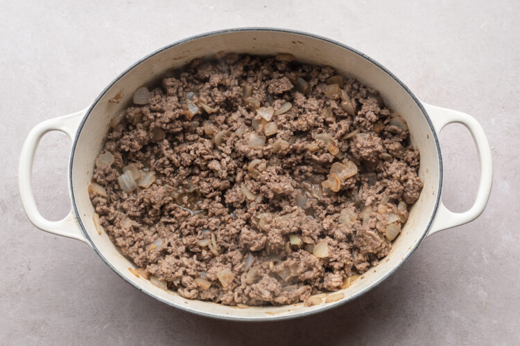 Overhead view of crumbled ground beef in a large chili pot with tender chopped onion and minced garlic on a white table.
