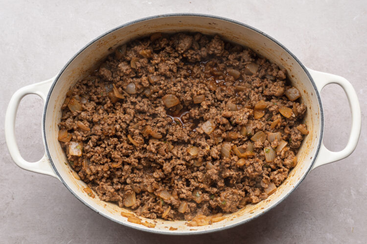 Overhead view of seasoned, crumbled ground beef in a large chili pot with tender chopped onion and minced garlic on a white table.