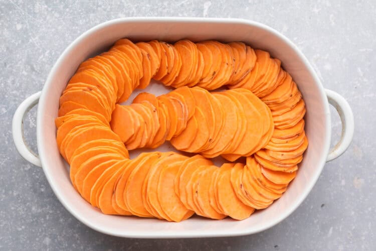 Overhead view of sweet potato slices arranged in a ring around the inside edge of an oval baking dish, with a row of slices in the center.