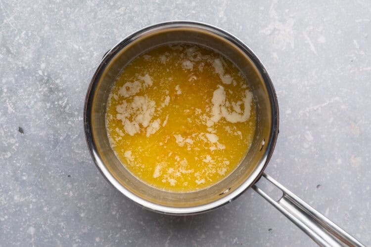 Overhead view of melted butter in a small saucepan on a neutral blue-gray background.