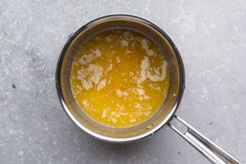 Overhead view of melted butter in a small saucepan on a neutral blue-gray background.