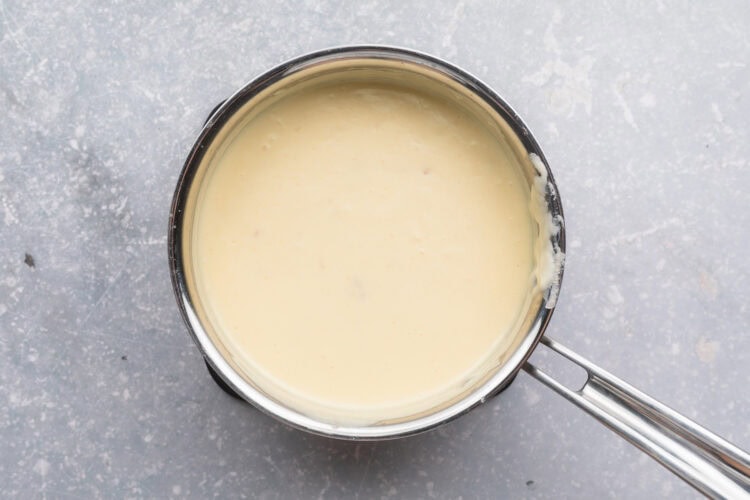 Overhead view of a small silver saucepan filled with a heavy cream sauce on a neutral blue-gray background.