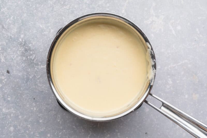 Overhead view of a small silver saucepan filled with a heavy cream sauce on a neutral blue-gray background.