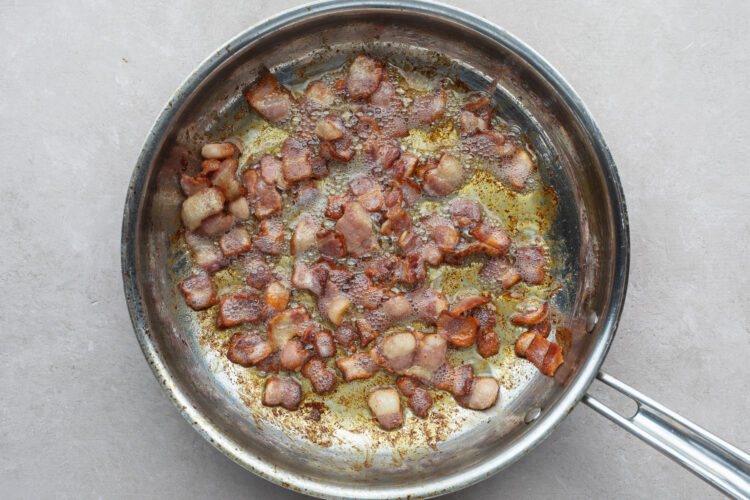 Overhead view of crispy diced bacon in a large silver skillet on a neutral countertop.