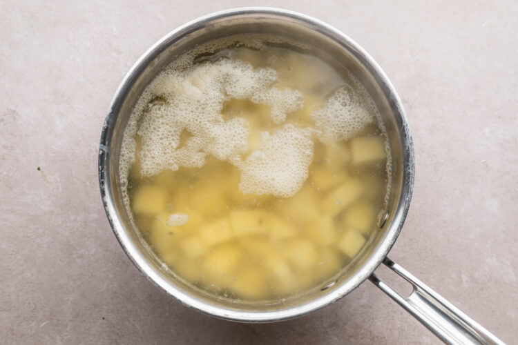 Overhead view of a silver pot holding boiling water, cubed potatoes, and salt on a neutral countertop.