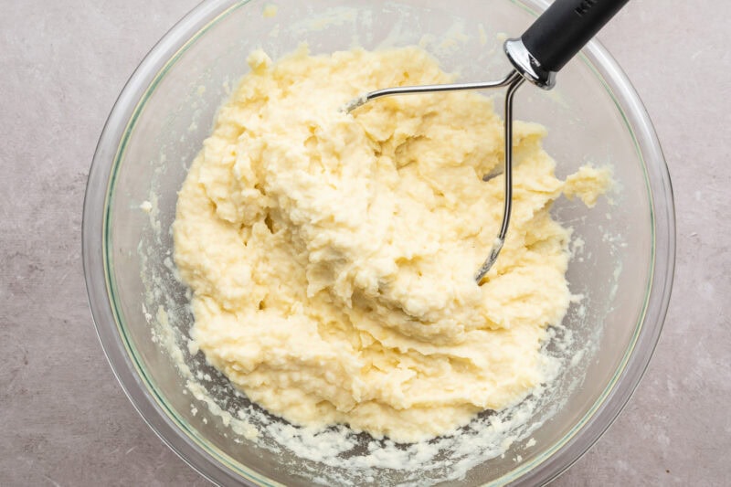 Overhead view of creamy mashed potatoes in a large glass mixing bowl with a potato masher resting on a neutral countertop.