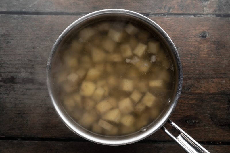 Overhead view of large silver pot holding cubed potatoes and cold salted water sitting on a wooden tabletop.