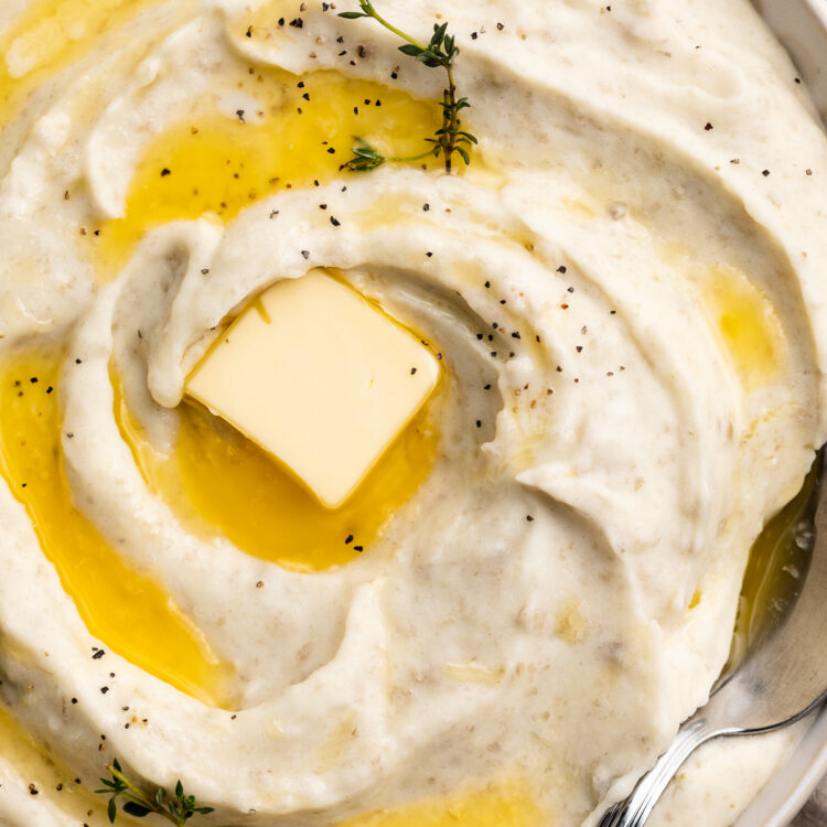 Close-up, overhead view of a large bowl of creamy whipped potatoes, swirled with melted butter and topped with a pat of butter and fresh herbs.