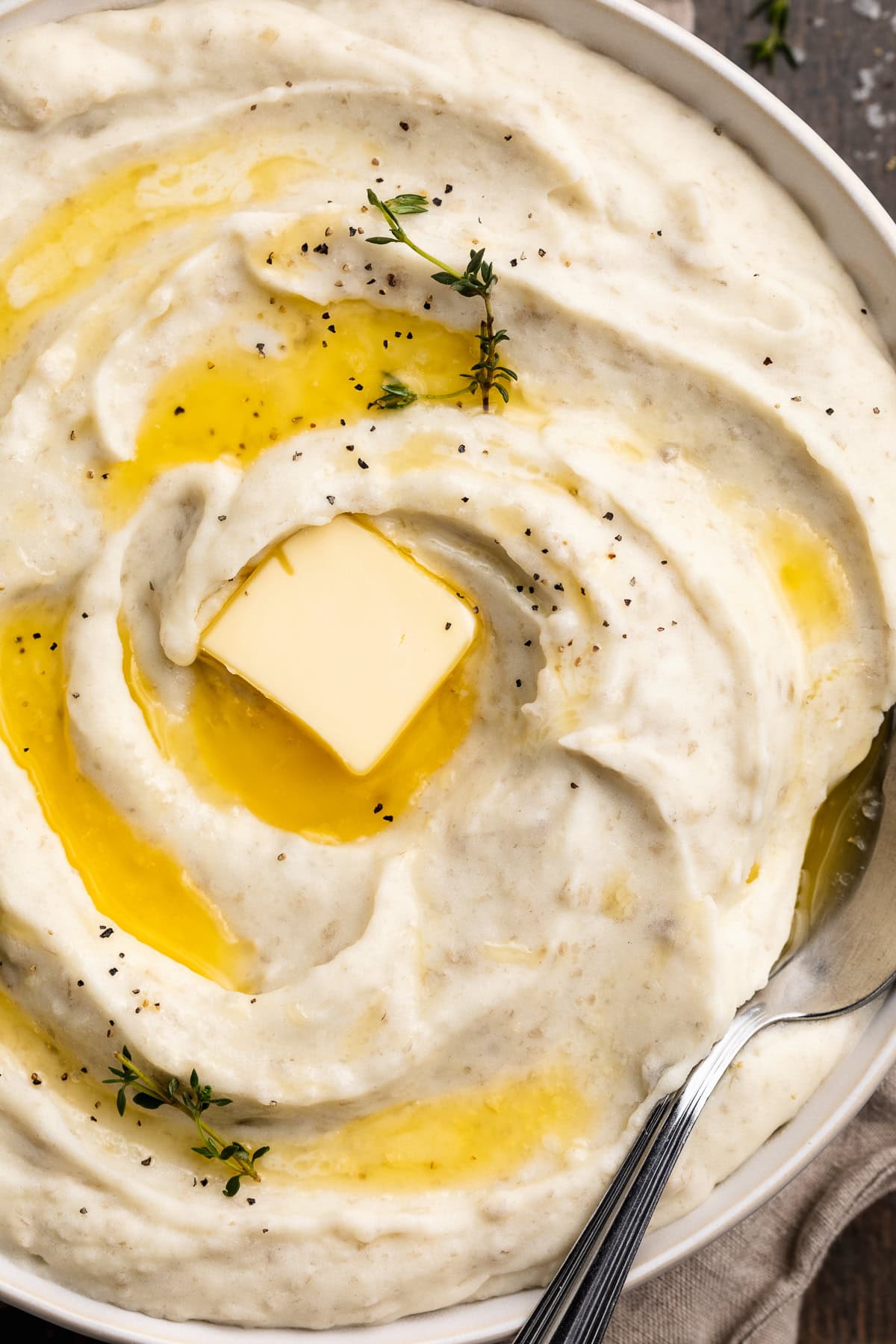Close-up, overhead view of a large bowl of creamy whipped potatoes, swirled with melted butter and topped with a pat of butter and fresh herbs.