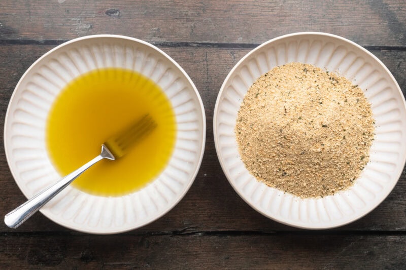 Overhead view of two shallow bowls, one containing an egg and oil wash and the other breadcrumbs.