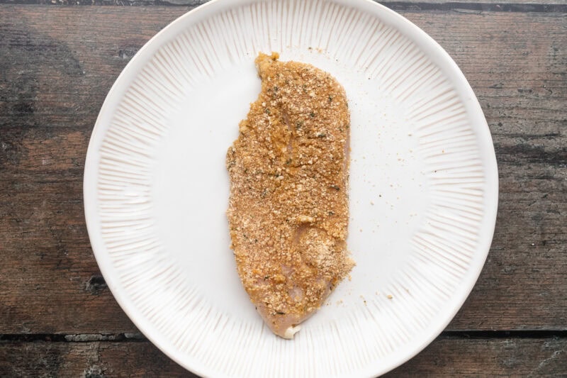 Breaded chicken cutlet resting on a large round white plate on a wooden table.