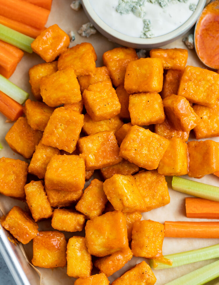 Squares of crispy orange-colored buffalo tofu on a sheet pan with celery sticks and carrot sticks next to a bowl of blue cheese dressing.