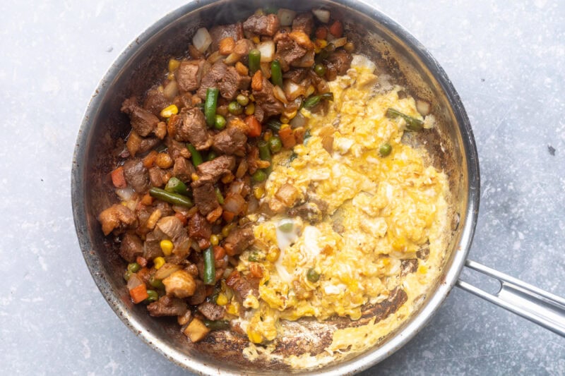 Duck breast, soy sauce, spices, and frozen mixed vegetables in a large silver skillet with half the pan holding scrambled eggs.
