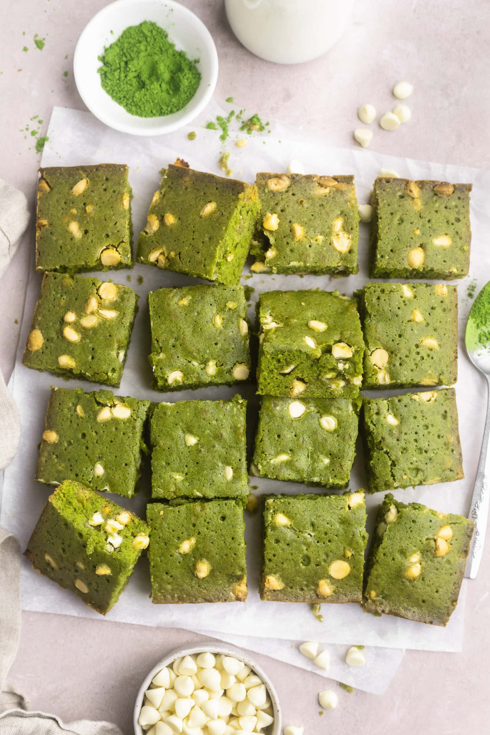 Squares of bright green matcha brownies peppered with white chocolate chips stacked up on parchment paper on a countertop.