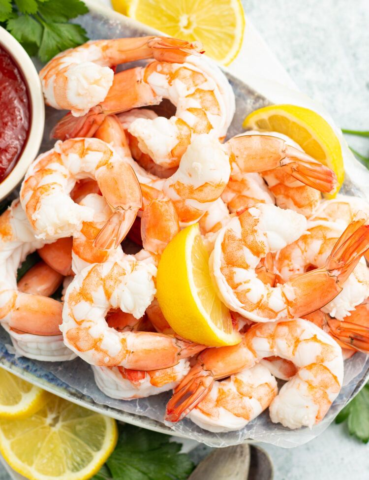 Angled, overhead photo of a tray holding poached shrimp on a bed of ice, with lemon wedges and sauce.